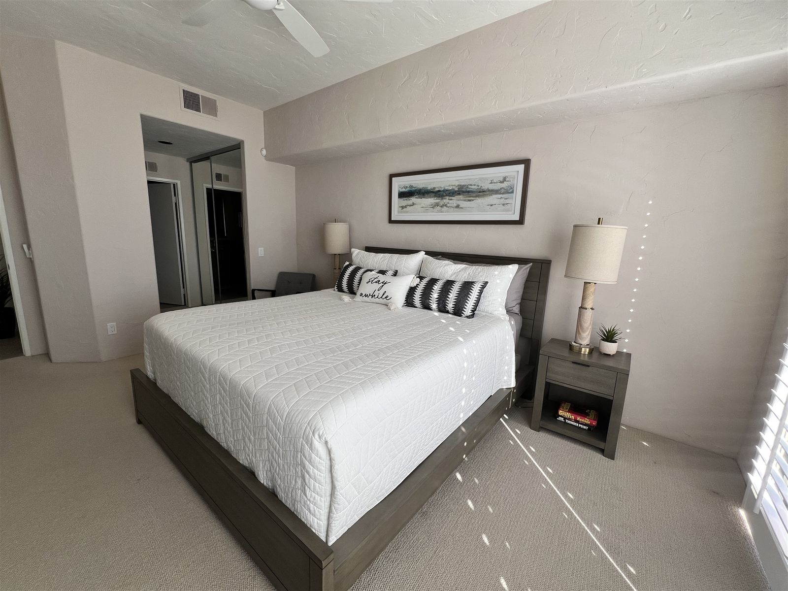 A hotel room with a large bed, two bedside lamps, a small table with a plant, and a framed picture on the wall.
