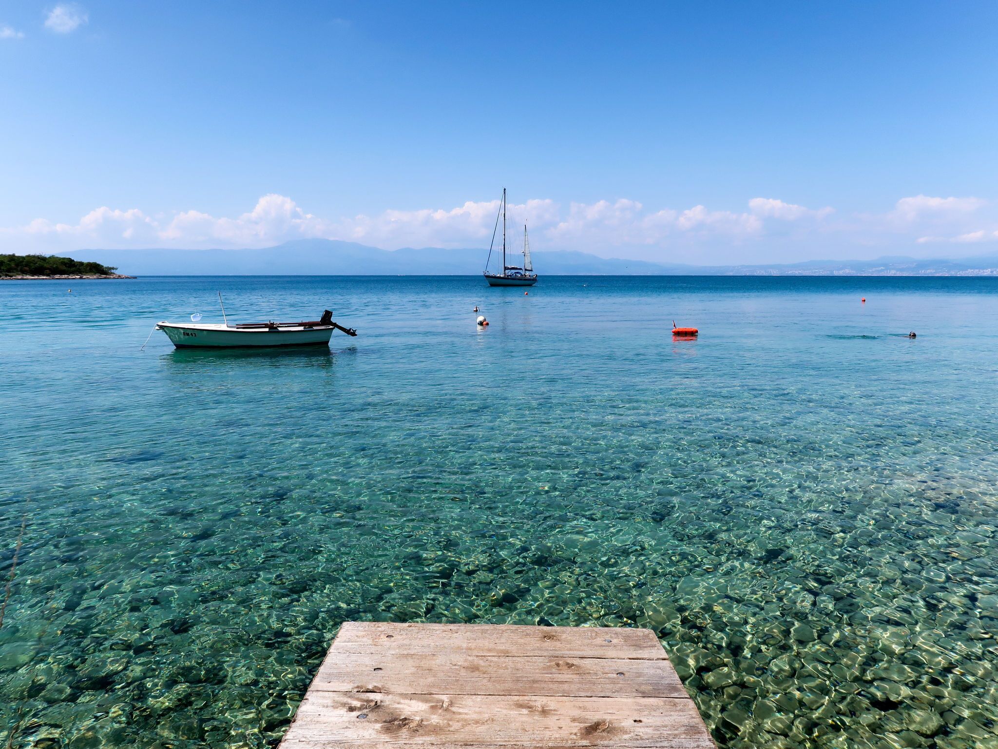 Eine Holzsteg erstreckt sich in ruhiges, seichtes Wasser, im Hintergrund liegen ein Boot und ein Segelboot vor Anker.