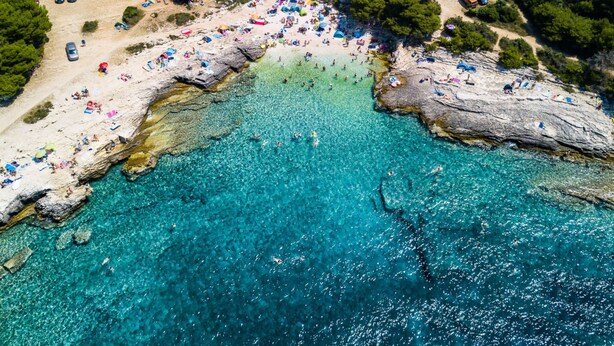 Oversigtsbillede af et kystområde med strand, klart turkis vand, klippeformationer og parkerede biler.