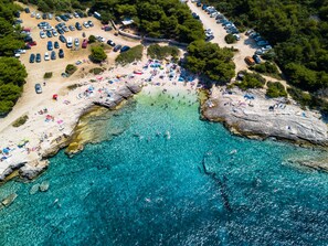 Luftaufnahme einer Küstenlandschaft mit Strand, klarem türkisblauem Wasser, Felsformationen und geparkten Autos.