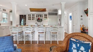 A spacious kitchen with white cabinetry, a central island with bar stools, and a dining area with a blue patterned sofa. - Pau Hana - Spacious 4-bedroom house in quaint Nantucket with AC (Nantucket)