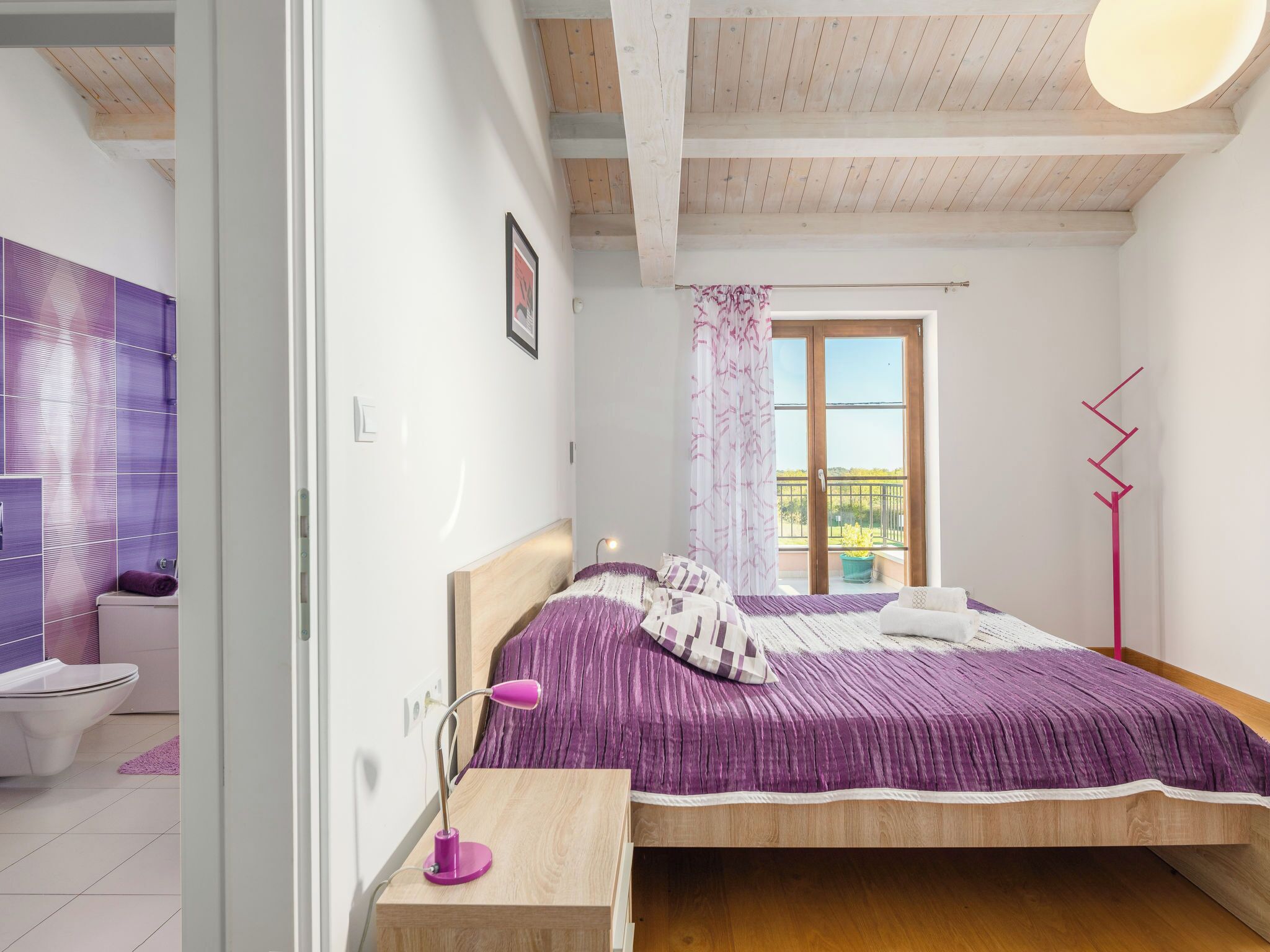 Une chambre à coucher avec un lit, une table de chevet avec une lampe, une fenêtre donnant sur un paysage agréable, et une salle de bain avec des toilettes et un lavabo.