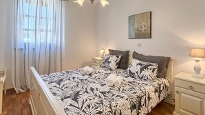 Een slaapkamer met een bed, gordijnen, een hanglamp, een nachtkastje met een lamp en een aan de muur hangend schilderij.