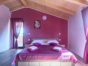 Un dormitorio con techo de madera, una cama con ropa de cama blanca y morada, un cuadro en la pared y una ventana con cortinas.