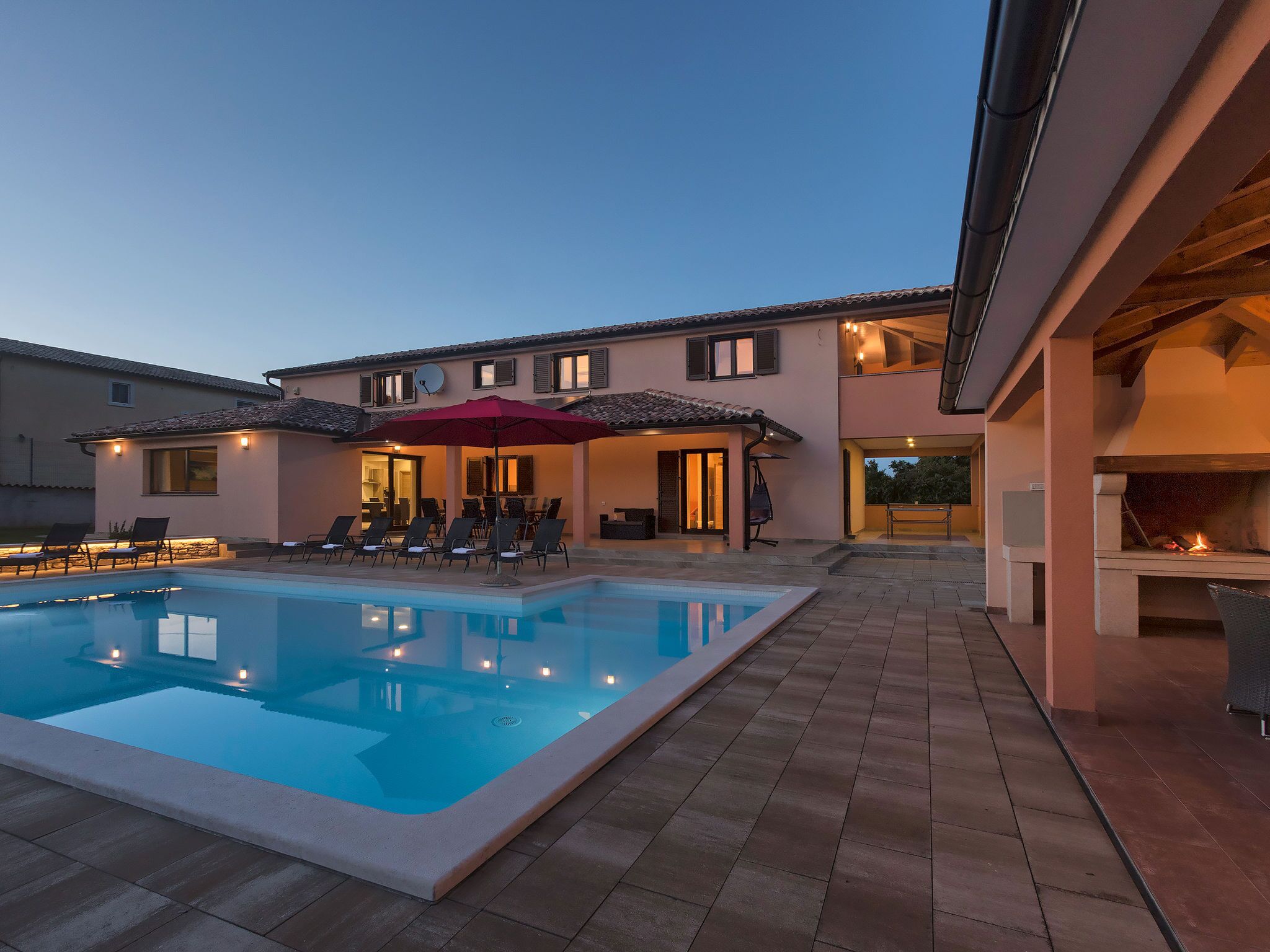 A swimming pool with lounge chairs and a covered seating area, a barbecue grill, and a two-story building with multiple windows.