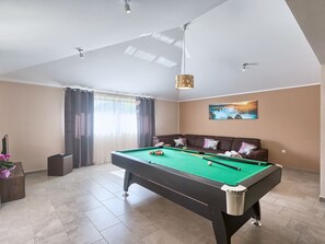 Une table de billard dans une pièce où se trouvent un canapé, un tableau au mur et une fenêtre avec des rideaux.