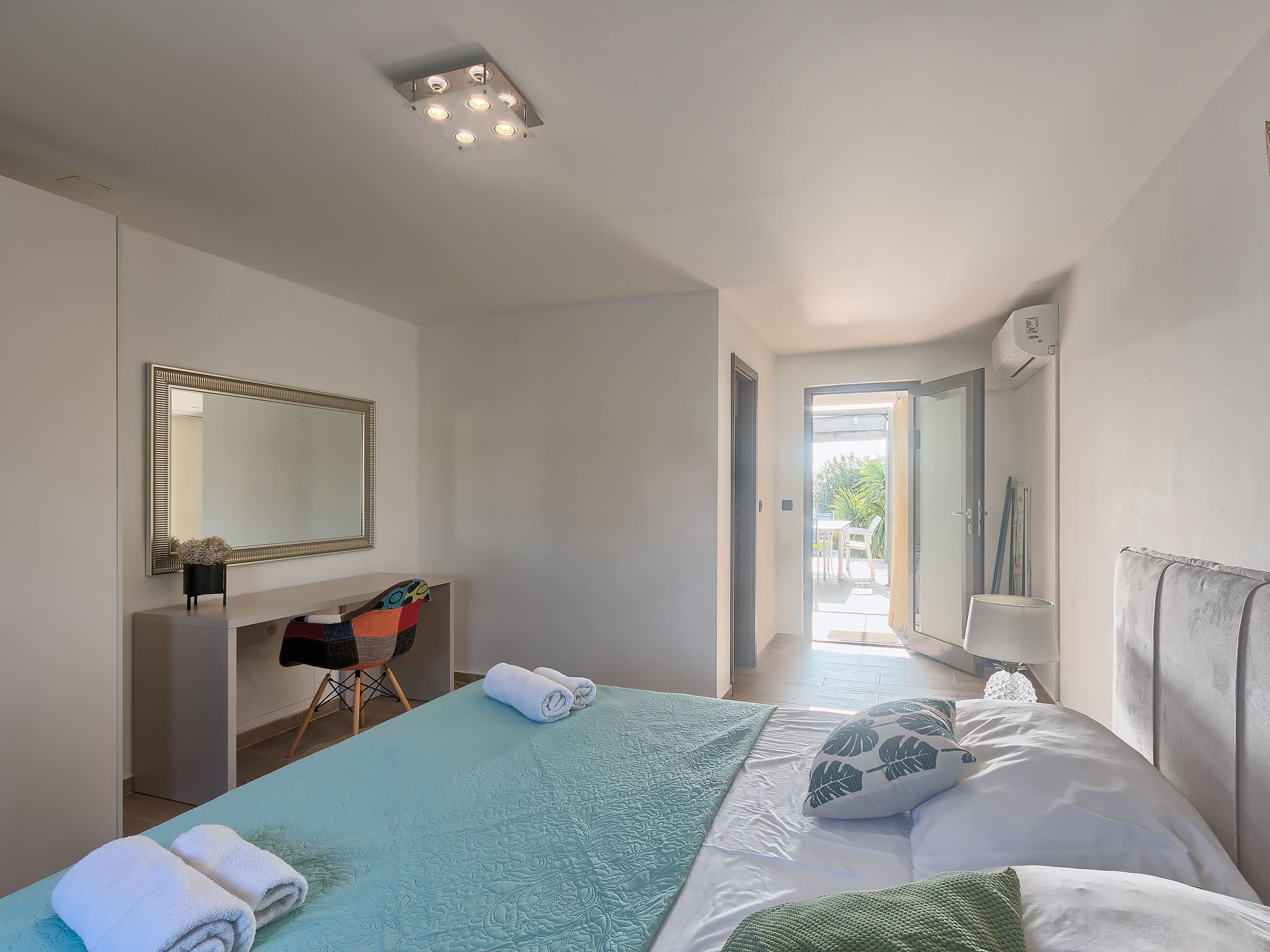 A bedroom with a bed, a mirror, a bedside table, and a view of the outdoors.