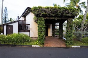 Ein Haus mit überdachter Veranda, einer kleinen Terrasse und einem Garten mit tropischen Pflanzen.