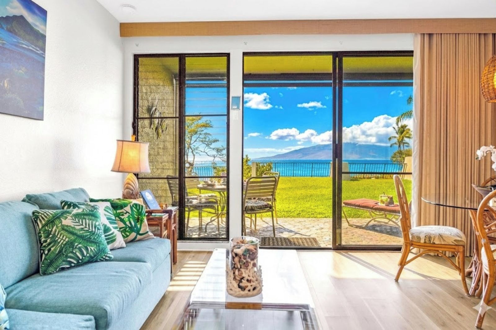 Sala de estar com sofá, mesa de centro e cadeiras, uma janela grande com vista para o oceano e uma montanha, e um quadro na parede.