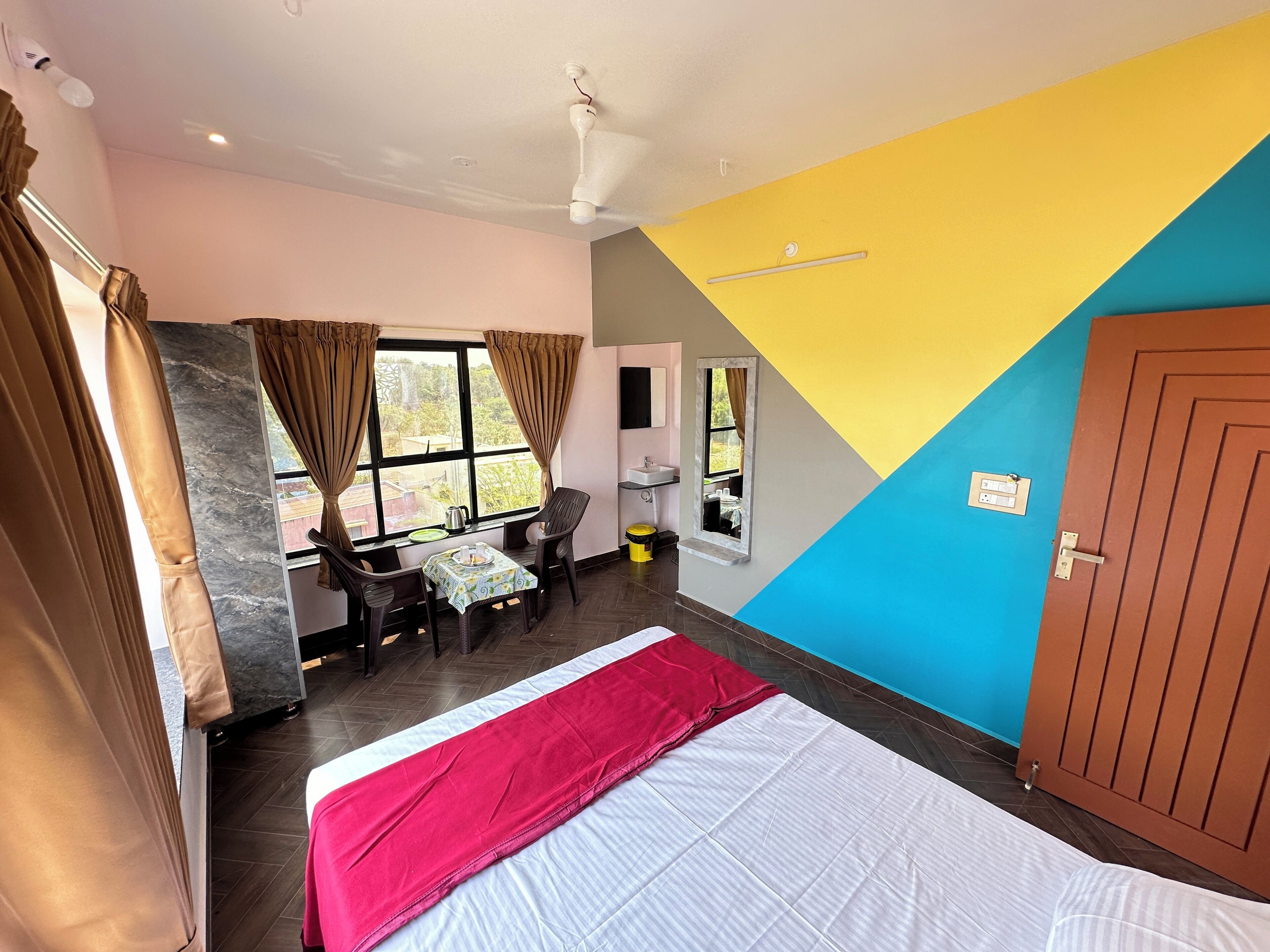 A room with a bed, a table, two chairs, a window with curtains, a sink, and a door.