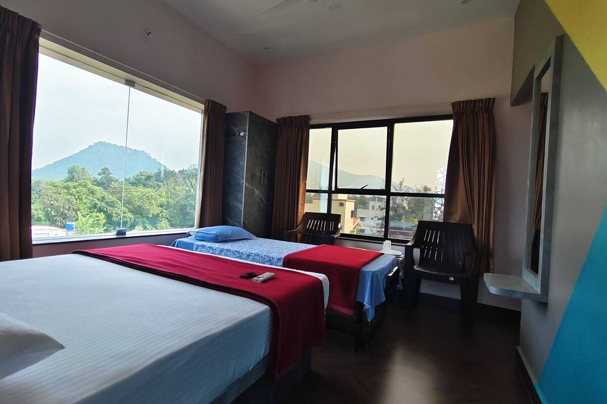 A hotel room with two beds, a desk, and a chair. There is a large window with a view of a mountain and greenery.