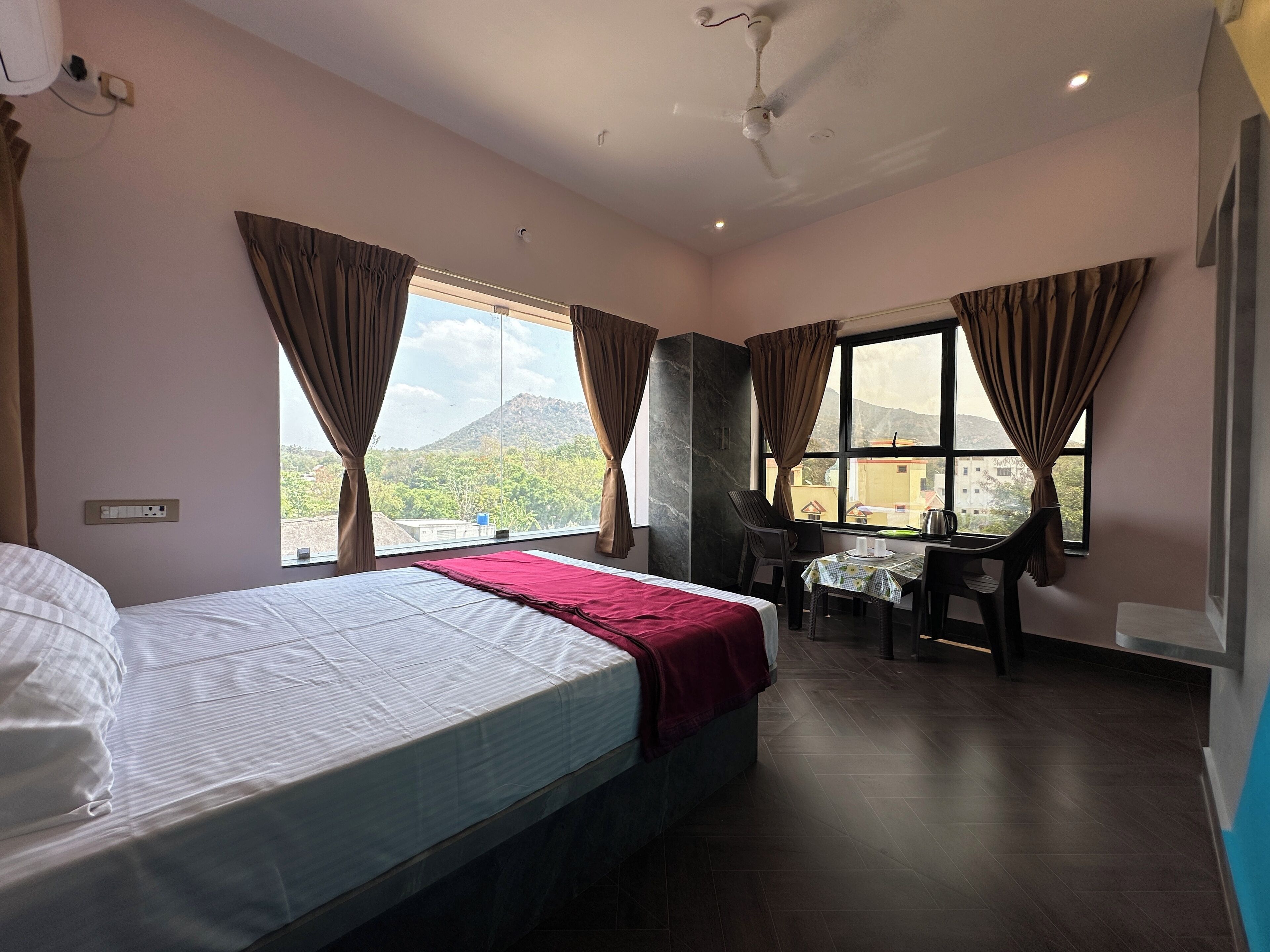 A hotel room with a bed, a desk, and a chair. There are windows with curtains, a ceiling fan, and a view of a mountain.