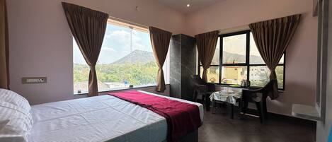 Quarto de hotel com cama, escrivaninha e cadeira. Janelas com cortinas, ventoinha no teto e vista para uma montanha.