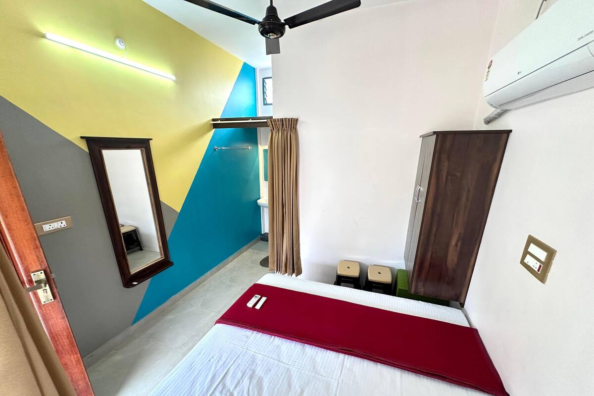 A hotel room with a bed, a mirror, a ceiling fan, a bathroom with a towel rack, and a wall-mounted air conditioner.