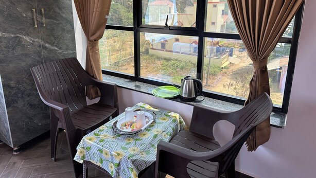 A room with a view of a mountainous landscape through large windows, a table with a floral tablecloth, two plastic chairs, and a kettle on the windowsill. - OM LINGESWARAN Home Stay Tiruvannamalai (Tiruvannamalai)