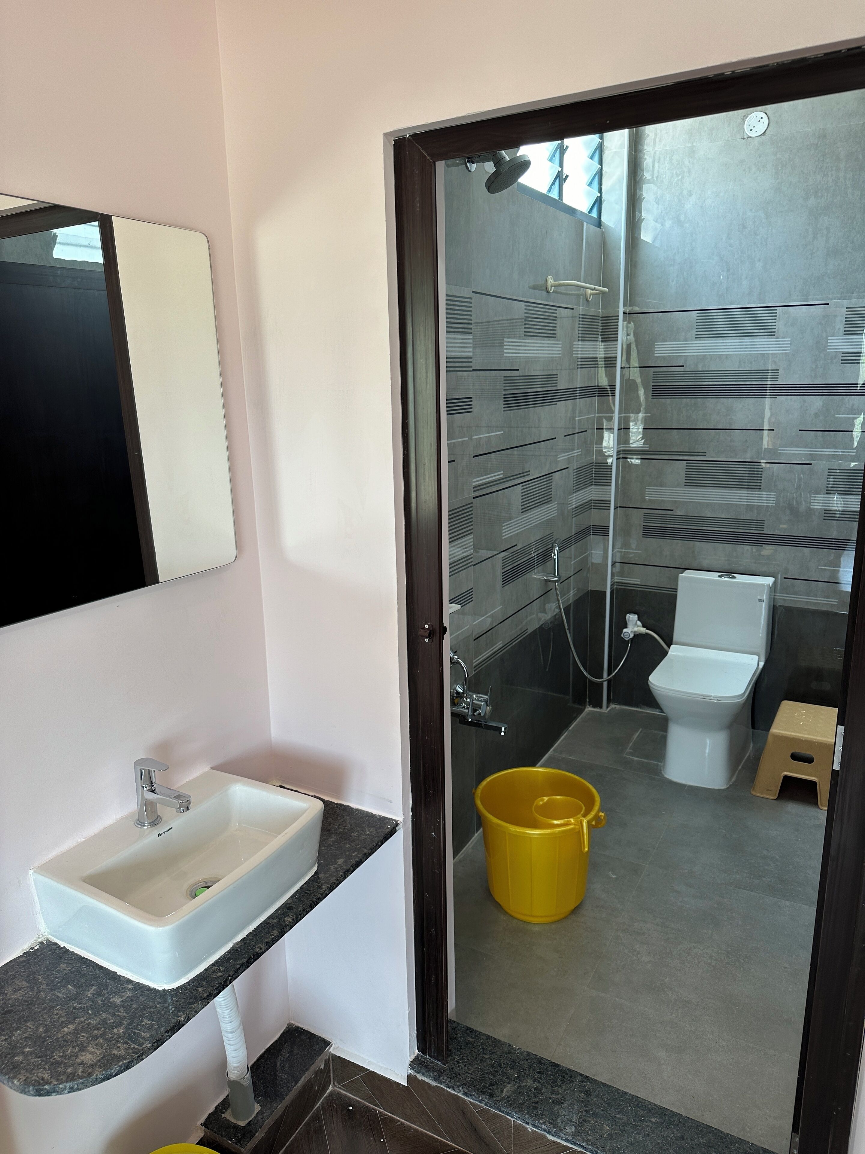 A bathroom with a white sink, a mirror, a yellow bucket, a toilet, and a shower area.