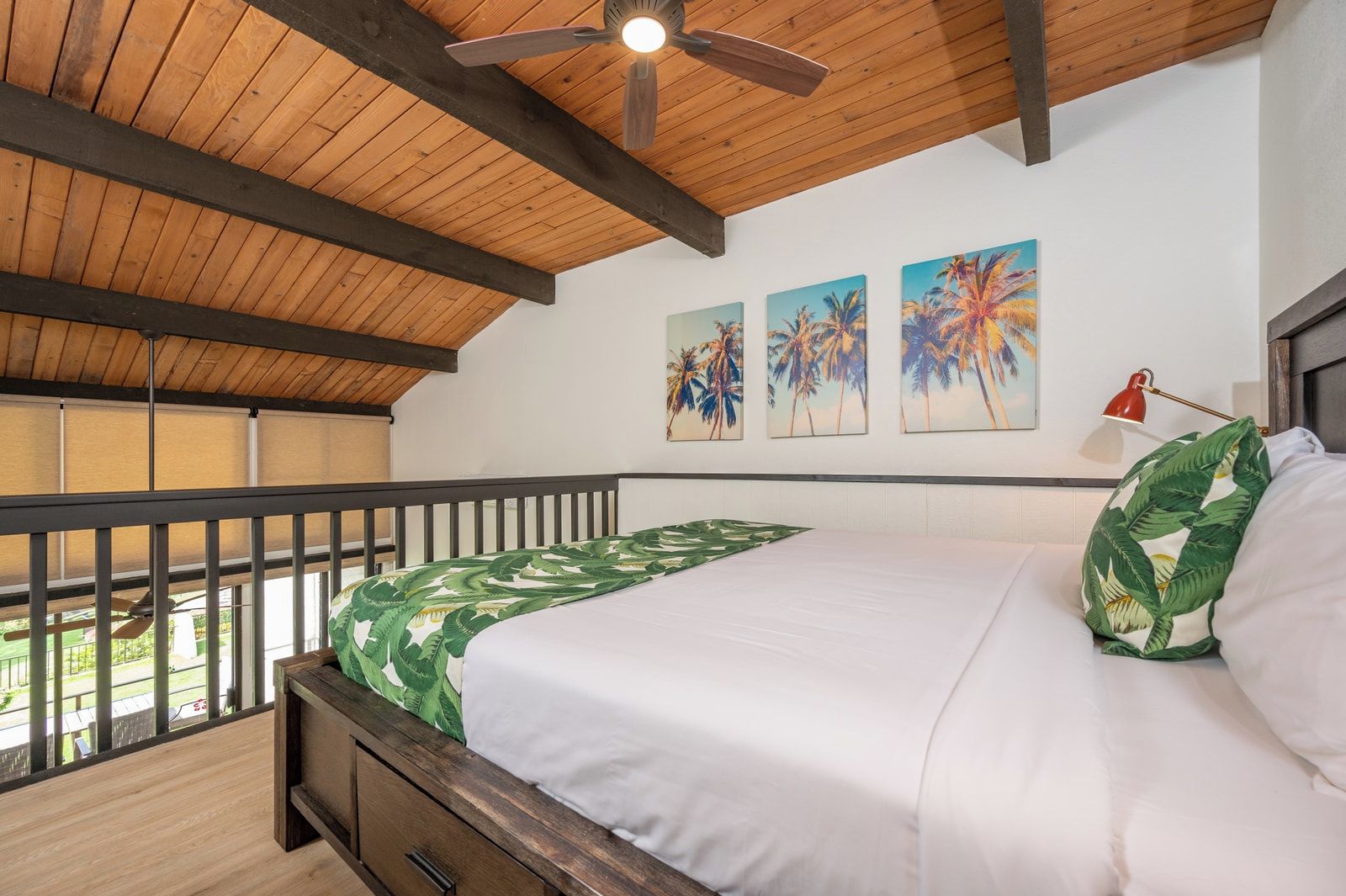 Ein Schlafzimmer mit Holzboden, einem Bett mit Bettdecke im Grün-Blatt-Muster, einer roten Schreibtischlampe und drei Palmenbildern an der Wand.