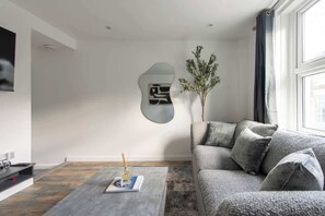 A modern living room with a grey sofa, a coffee table with a book and a vase, and a wall-mounted abstract art piece. - 4-Bed Maidstone House Sleeps 8 | Near East Station (Maidstone)