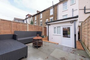 A patio with a sofa and a table, a wooden fence, and a building with a door. - 4-Bed Maidstone House Sleeps 8 | Near East Station (Maidstone)