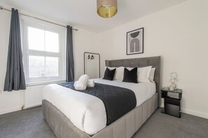 Une chambre à coucher comprenant un lit, une table de chevet, une fenêtre avec des rideaux, un radiateur et une œuvre d’art accrochée au mur.