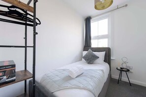 A single bed with a grey headboard, a bedside table with a lamp, a window with curtains, and a bunk bed structure to the left. - 4-Bed Maidstone House Sleeps 8 | Near East Station (Maidstone)
