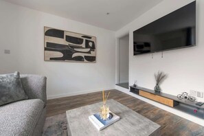 Un salon moderne avec un canapé gris, une table basse sur laquelle se trouvent un livre et un vase, un grand tableau abstrait accroché au mur, un téléviseur à écran plat et une console en bois agrémentée d’objets décoratifs.