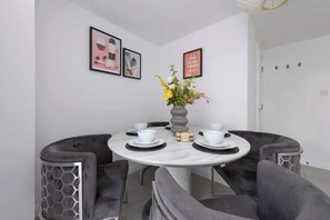 A dining area with a round table, grey upholstered chairs, a vase with flowers, and framed pictures on the wall. - 4-Bed Maidstone House Sleeps 8 | Near East Station (Maidstone)