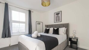Une chambre à coucher comprenant un lit, une table de chevet, une fenêtre avec des rideaux et une œuvre d’art accrochée au mur.