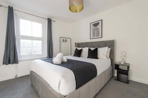 A bedroom with a bed, a bedside table, a window with curtains, and a wall-mounted artwork.
