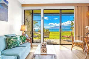 A living room with a sofa, coffee table, and chairs, a large window with a view of the ocean and a mountain, and a painting on the wall. - Napili Point | Oceanfront 4 BR, Sleeps 14 | Car Incl. w/ 6+ Nights | NAP ML-3622 by KBM (Lahaina)