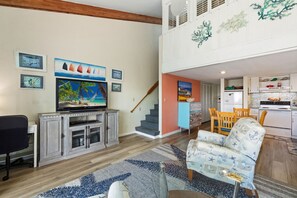 A living room with a television, a dining area with a table and chairs, a kitchen with appliances, and a staircase leading to an upper level. - A Wave From It All at Park Place (Cedar Key)