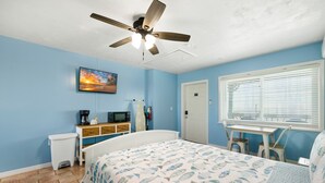 Un dormitorio con cama, ventilador de techo, televisor, microondas, cafetera, una tabla de surf y una ventana con persianas.