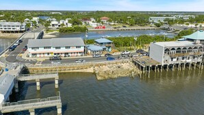 Un pueblo costero con un muelle que se adentra en el agua, una carretera con vehículos y edificios con comercios.