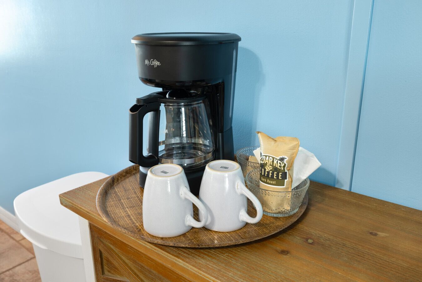 A coffee maker, two white mugs, and a bag of coffee on a wooden tray.