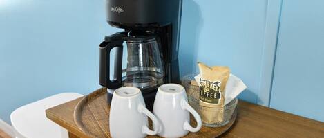 A coffee maker, two white mugs, and a bag of coffee on a wooden tray.