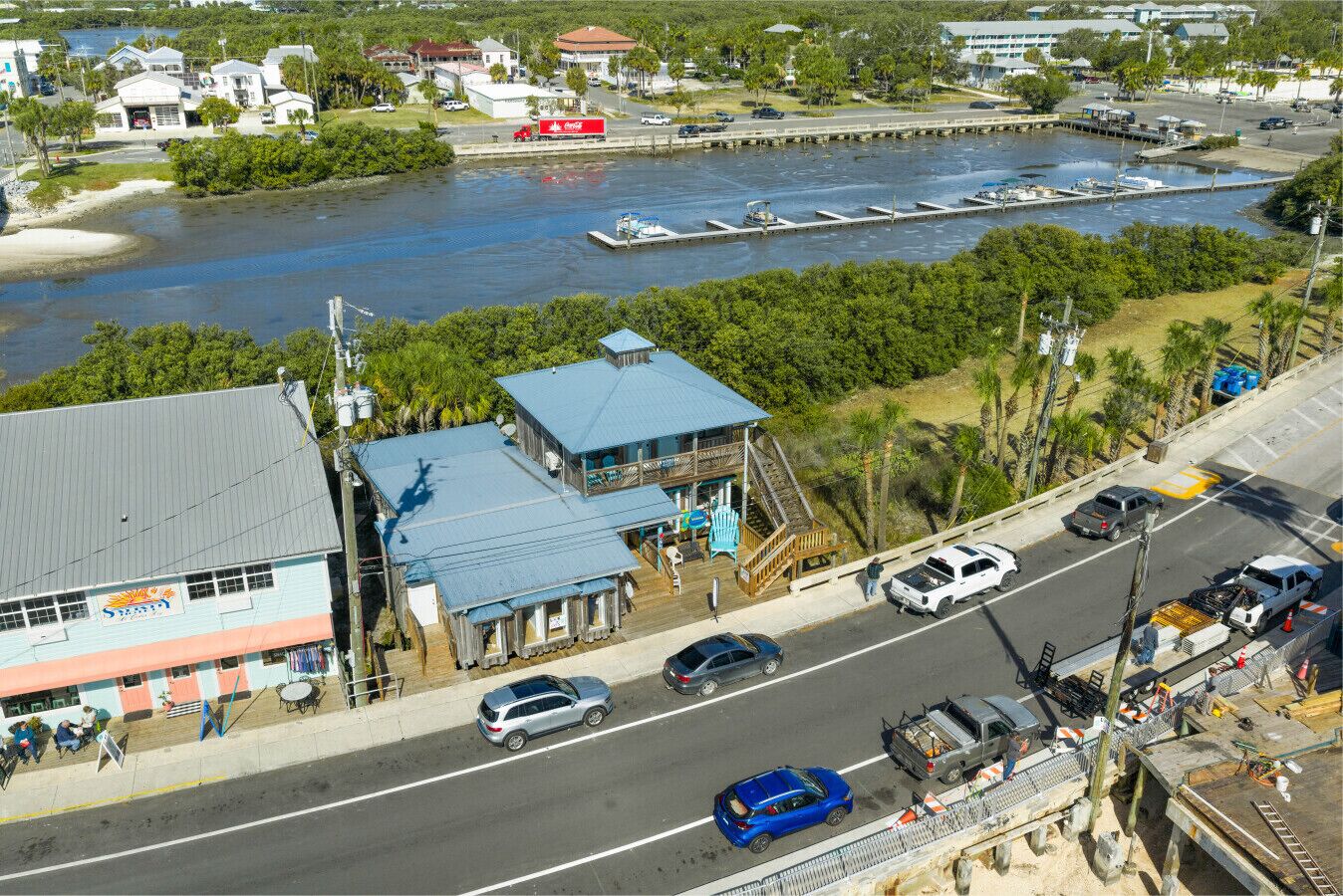 A coastal town with a dock, a restaurant, and a road with vehicles.