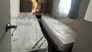 Une petite pièce, de taille modeste, avec deux lits recouverts de plastique protecteur, une tête de lit en bois, une table de chevet et une fenêtre avec des rideaux.
