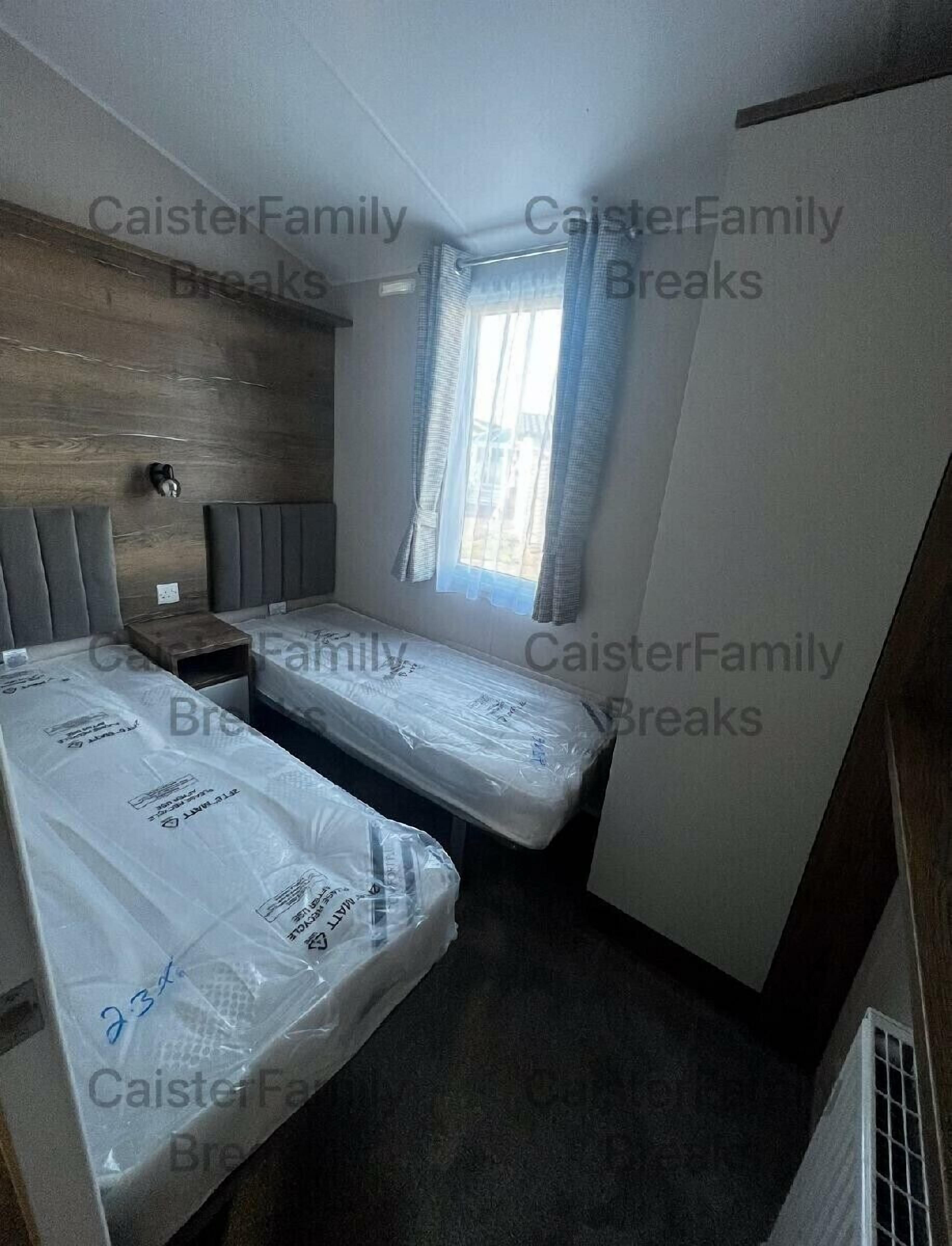 A compact room with two beds, a window with curtains, a wall-mounted light, and a wooden panel on the left.