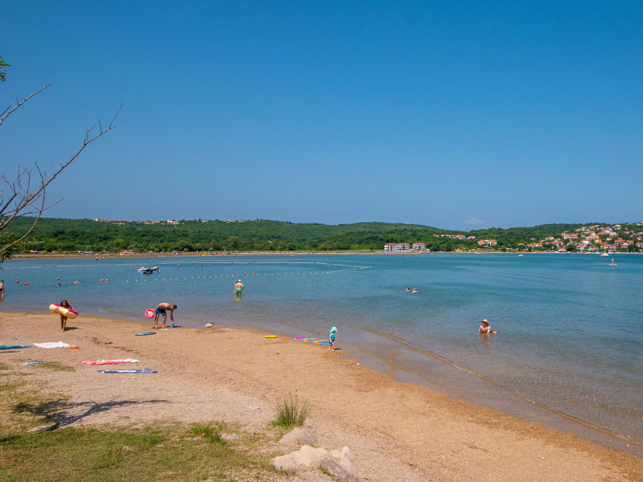 A beach with clear blue water, people swimming and sunbathing, and buildings in the background.