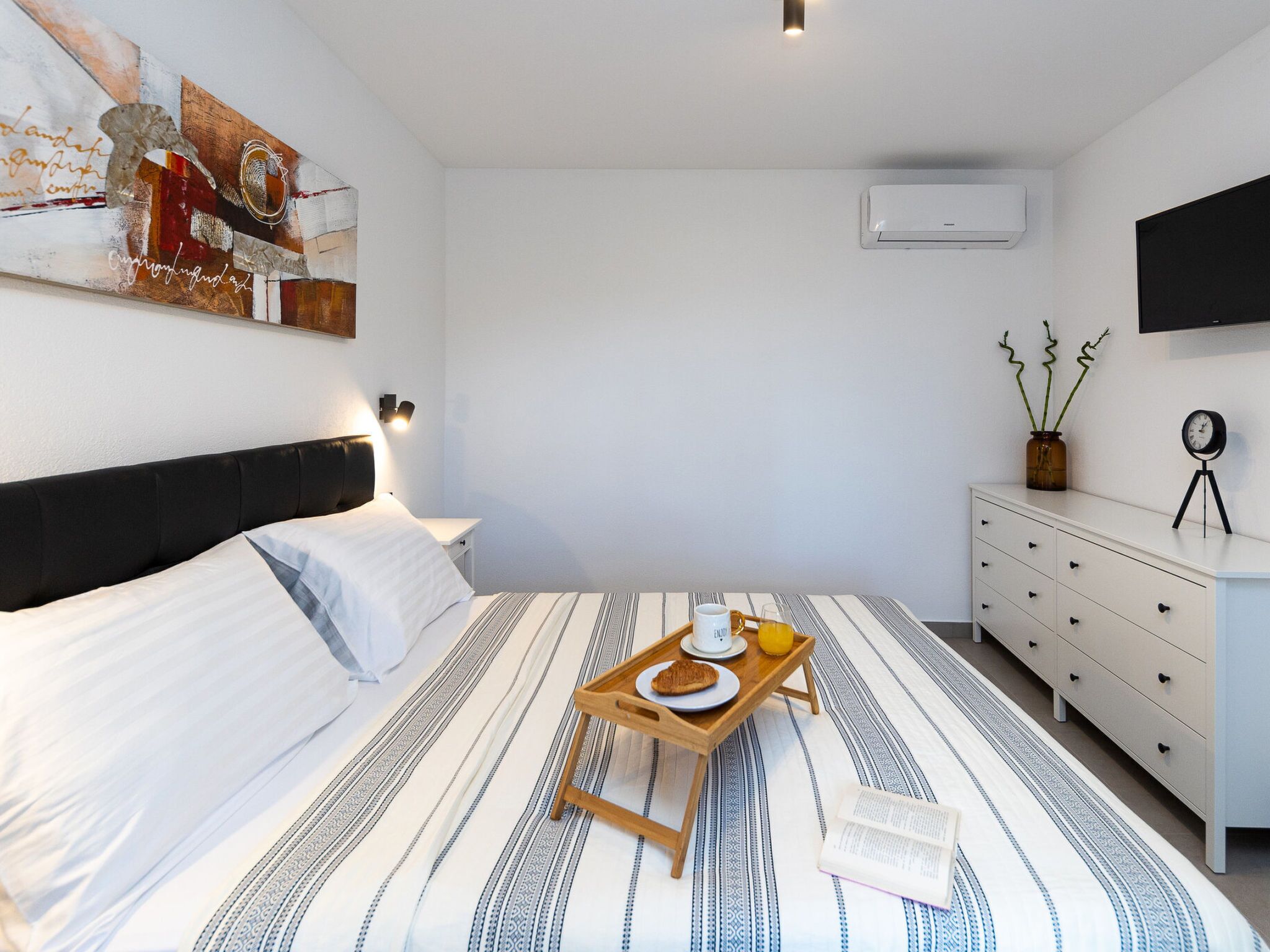 Ein Schlafzimmer mit Bett, Nachttisch, einem Tablett mit Frühstücksangeboten, einer Uhr und einem Flachbildfernseher an der Wand.