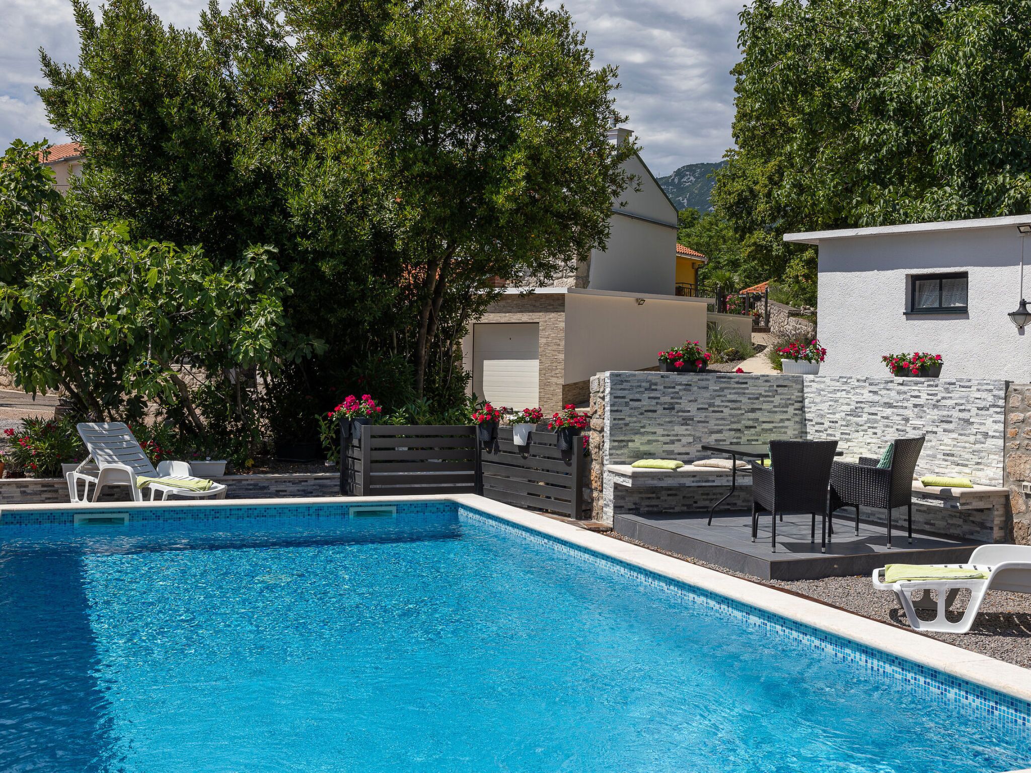 Ein Schwimmbecken mit Liegestühlen und einem Tisch, umgeben von Grünflächen und einer Steinmauer.