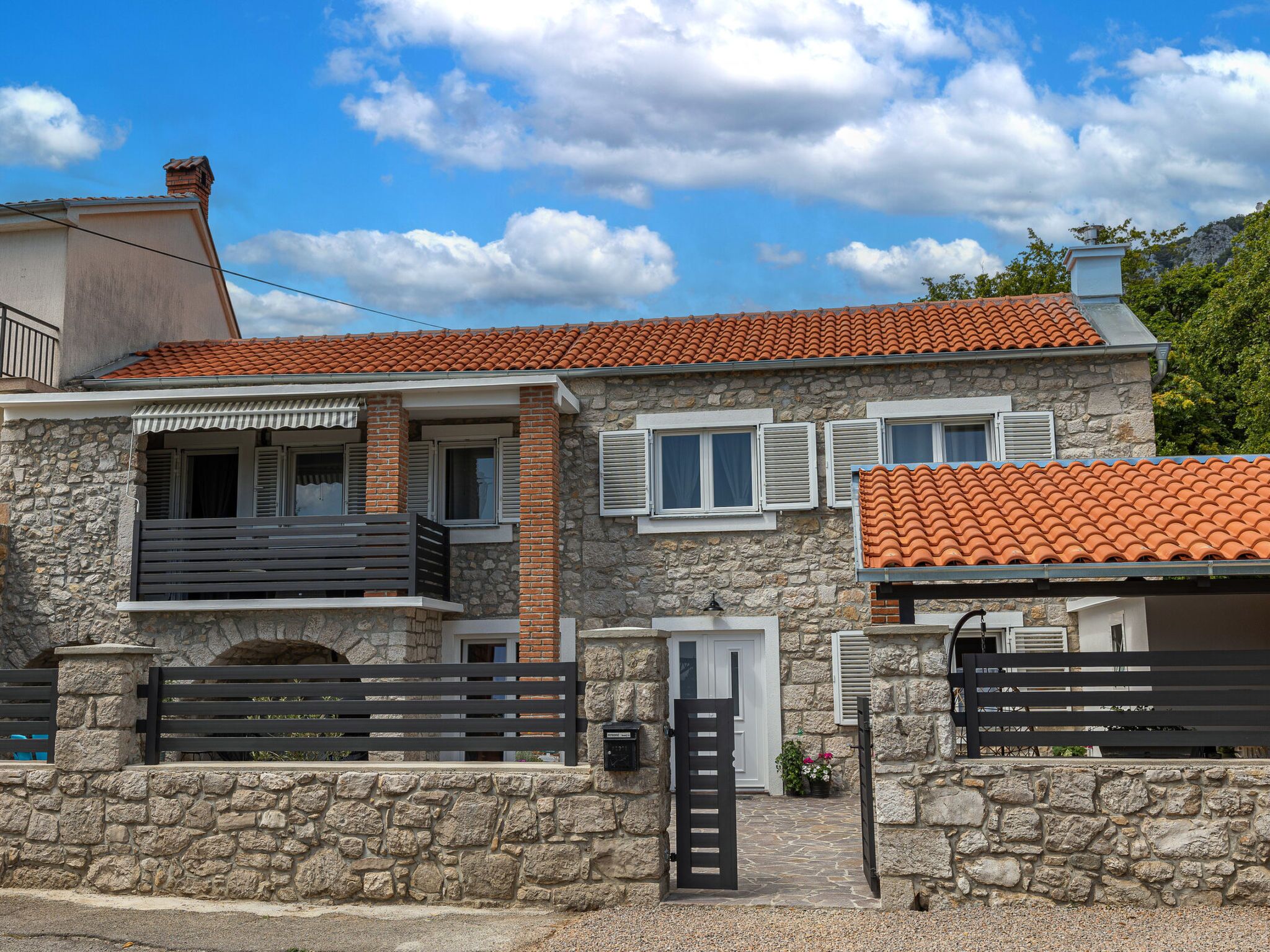 A two-story stone house with a tiled roof, a balcony, and a gated entrance.