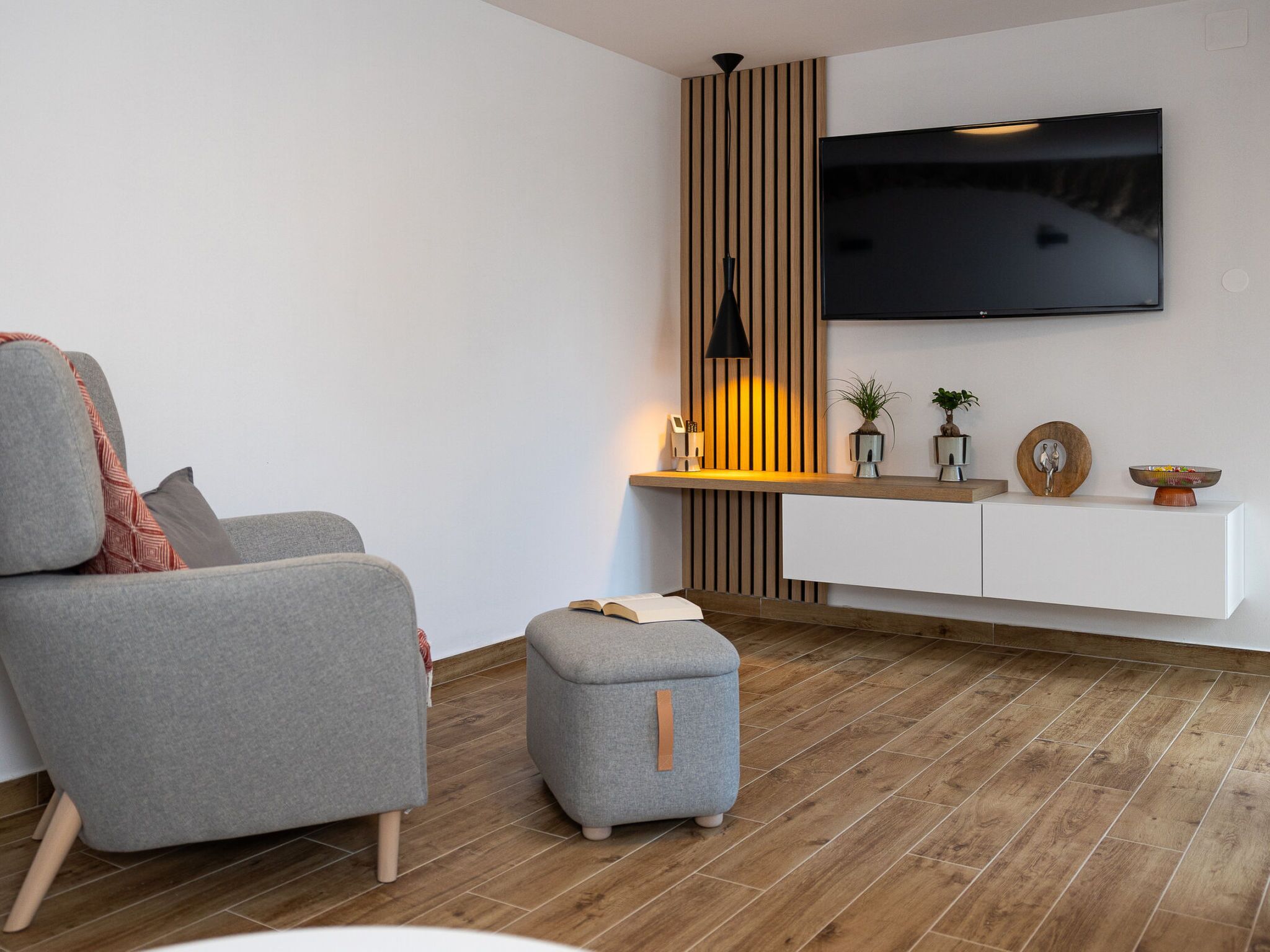 Een moderne woonkamer met een grijze bank, een klein poefje, een houten accentmuur, een wandgemonteerde televisie en een minimalistische dressoir.
