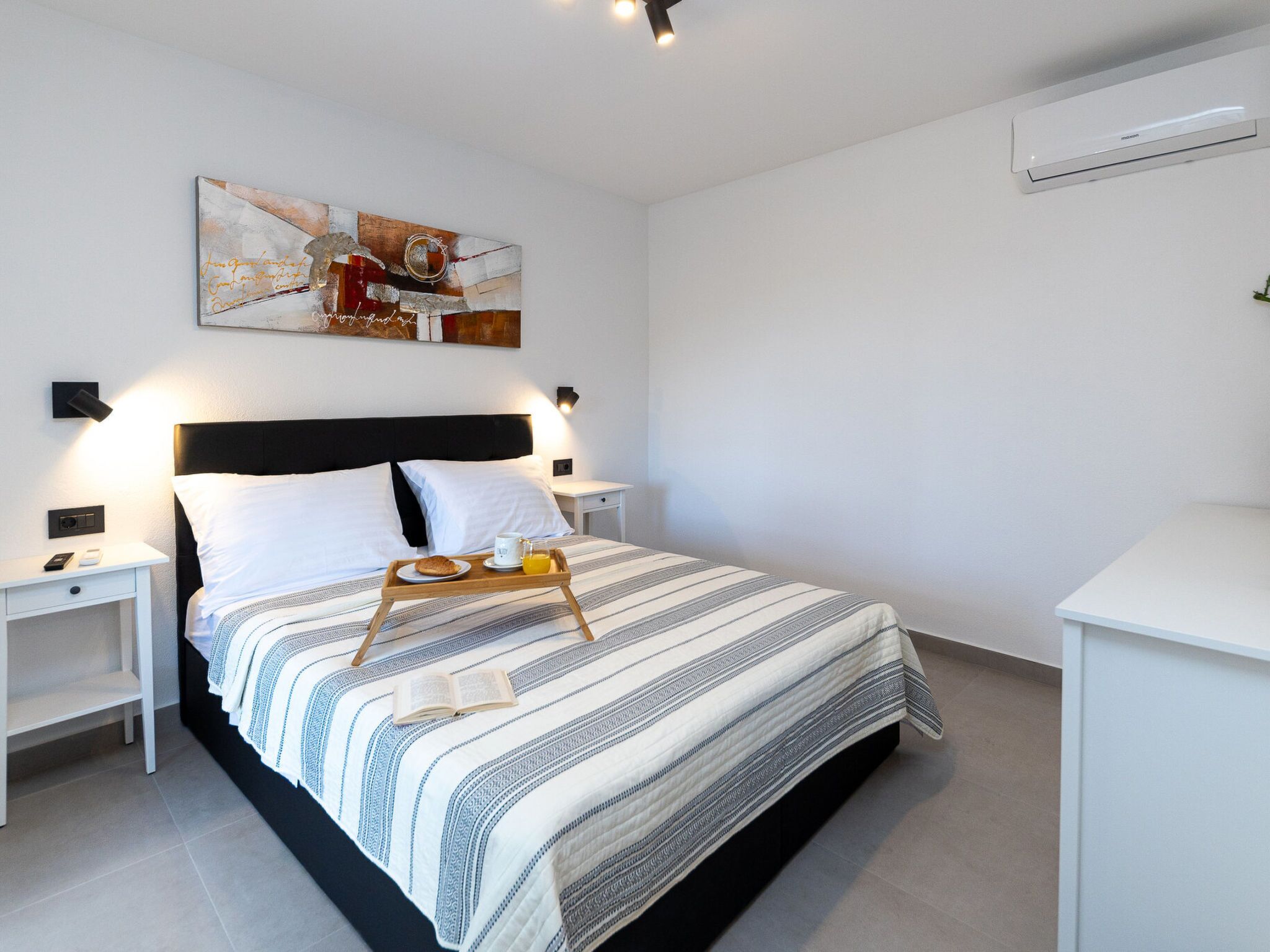 A modern hotel room with a bed, bedside tables, a nightstand, a bedside lamp, a tray with breakfast items, and a painting on the wall.