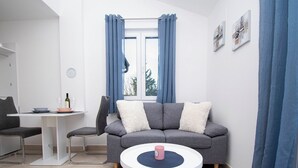 Ein modernes Wohnzimmer mit einem grauen Sofa, einem runden Couchtisch, einem Esstisch mit Stühlen und einem Fenster mit blauen Vorhängen.