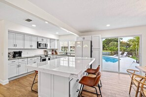 A modern kitchen with white cabinetry, a central island, and a view of a backyard swimming pool. - Waterfront Oasis | Heated Pool, MiniGolf, TikiHut (Lake Worth)