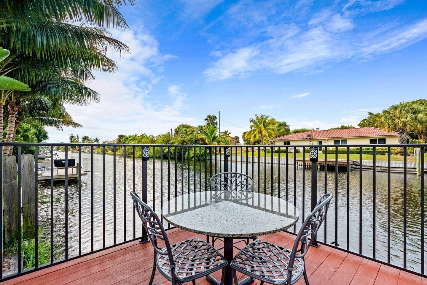 A balcony with a table and chairs, overlooking a body of water with a fence and palm trees.