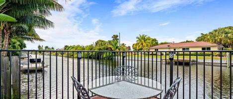 A balcony with a table and chairs, overlooking a body of water with a fence and palm trees.