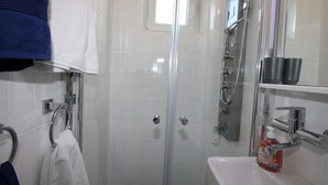 Kylpyhuone, jossa on lasiseinäinen suihkukaappi, valkoinen pesuallas ja hylly, jossa on hygieniatuotteita.