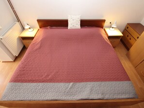 A neatly made bed with a red and grey quilt, wooden headboard, bedside tables with lamps, and a radiator. - Apartment Sasa A2 / HR-11069-03 by Interhome (Dracevac)